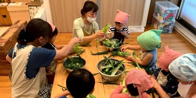 トウモロコシの皮をむく保育士と子供たち
