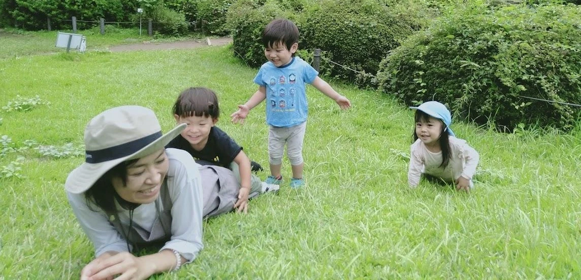 公園で寝転ぶ保育士と遊ぶ子供たち