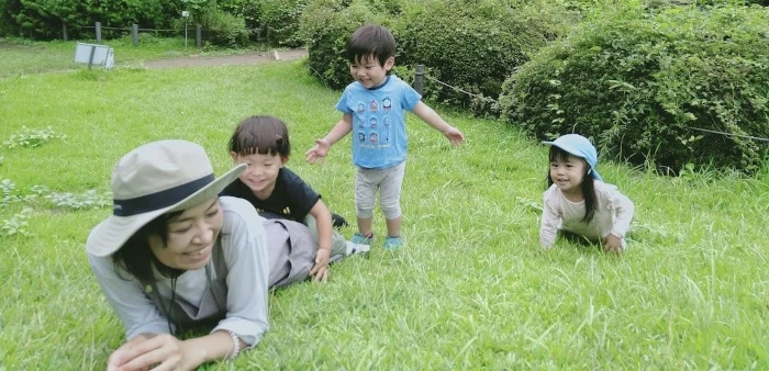 公園で寝転ぶ保育士と遊ぶ子供たち